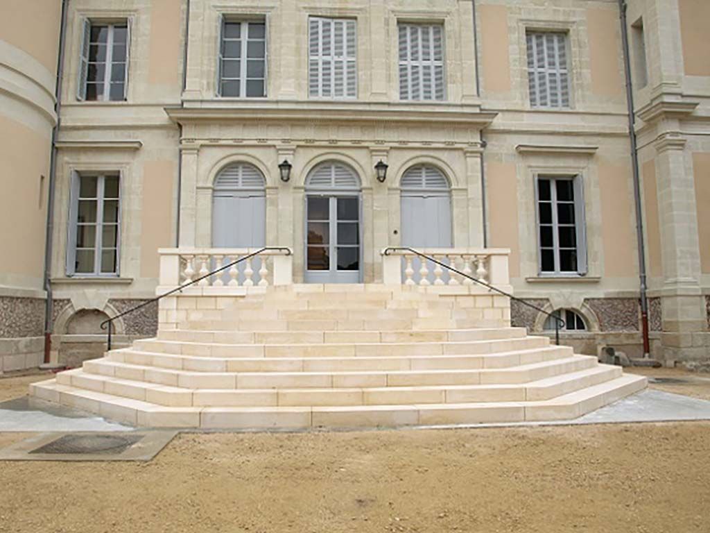 Un escalier en pierre mène à l'entrée d'un bâtiment symétrique de couleur claire, percé de nombreuses fenêtres.