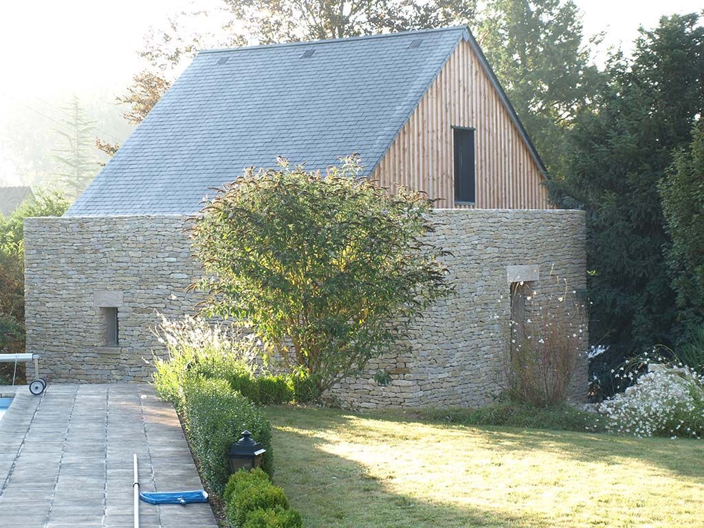 Un bâtiment en pierre avec un toit en ardoise et un pignon en bois, situé dans une cour arrière ensoleillée, à côté d'une terrasse en pierre et d'un petit arbre.