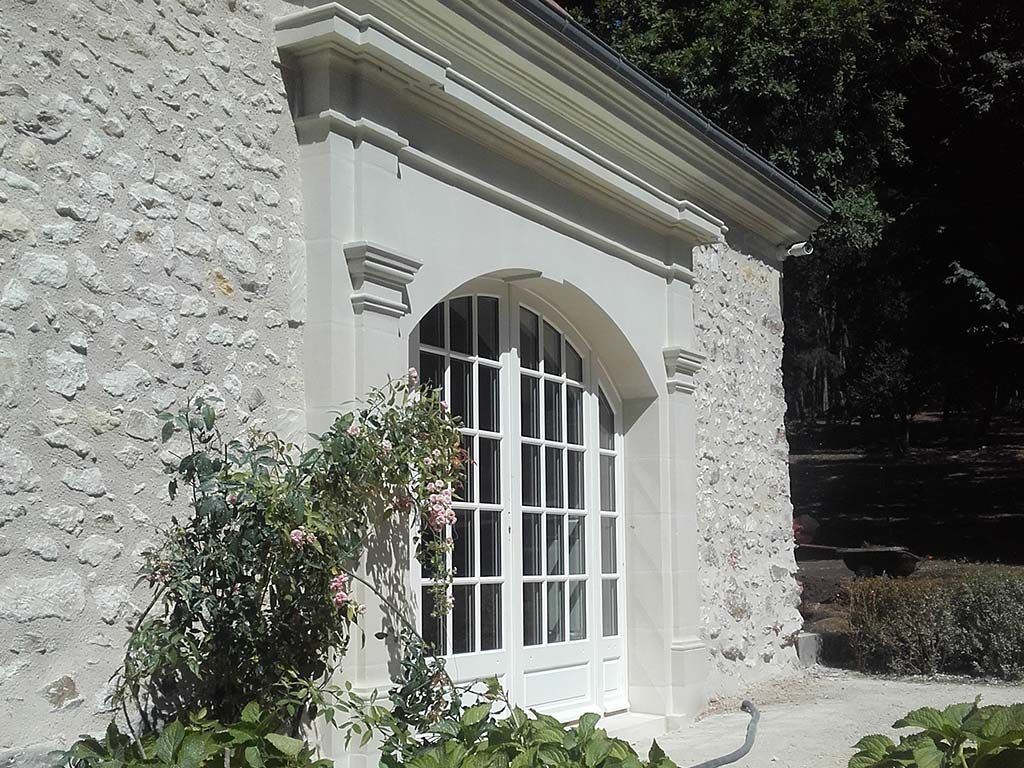 Une porte-fenêtre cintrée blanche, encadrée de pierre, sur un mur de pierre claire à l'aspect brut, avec un petit arbuste au premier plan.