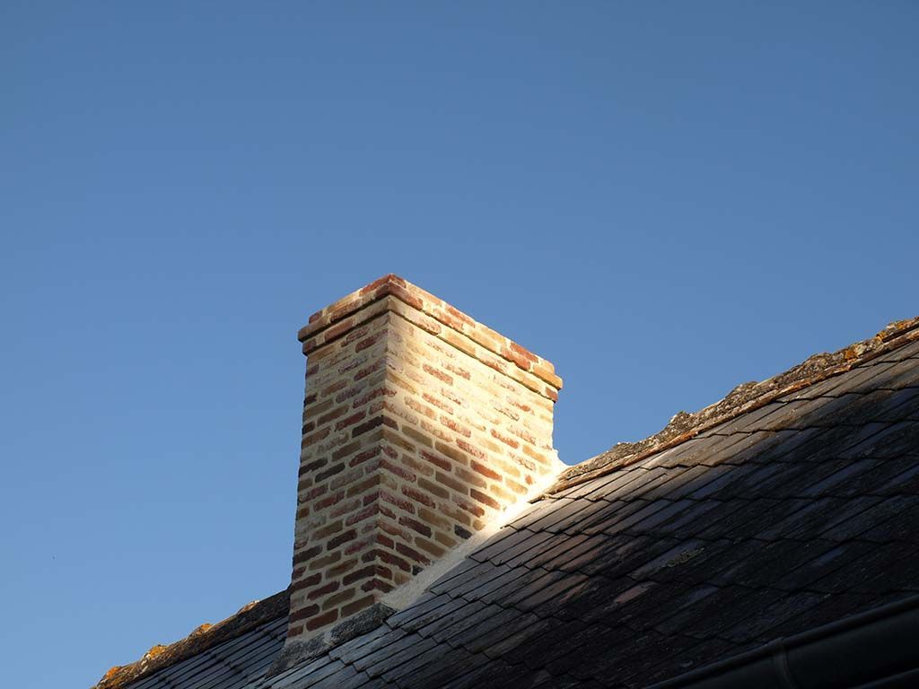 Une cheminée en briques se dresse sur un toit sombre et texturé, se détachant sur un ciel d'un bleu limpide.