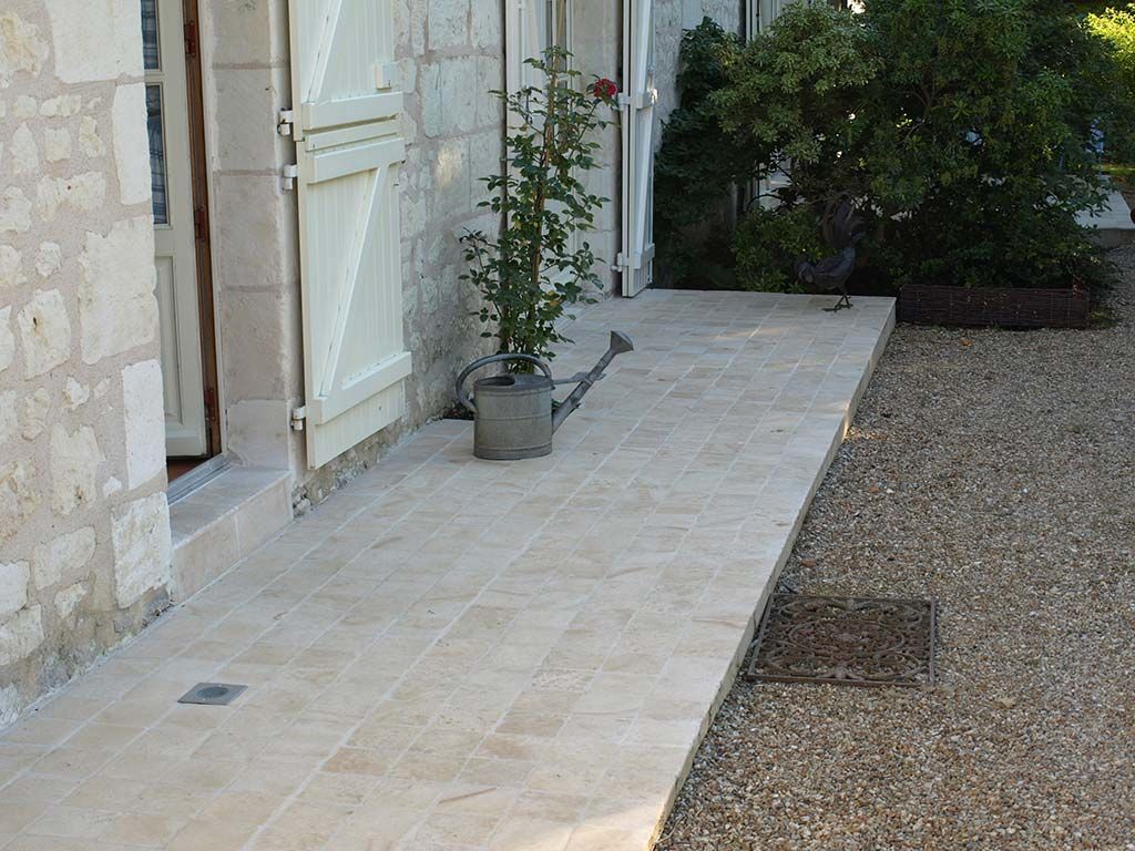 Une allée en pierre avec une plante en pot et un arrosoir, adjacente à un mur en pierre claire et à une zone de gravier.