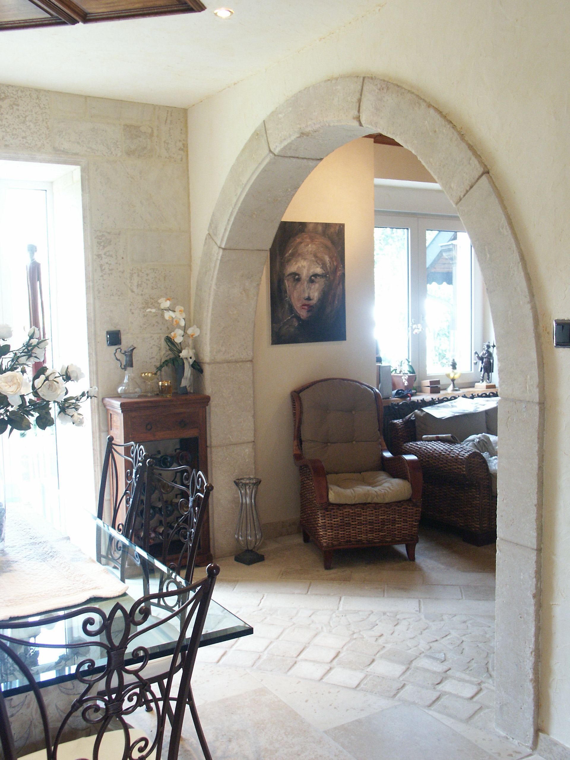 Une porte cintrée en pierre assure la transition entre un coin repas avec des meubles en métal et un espace salon avec un fauteuil.