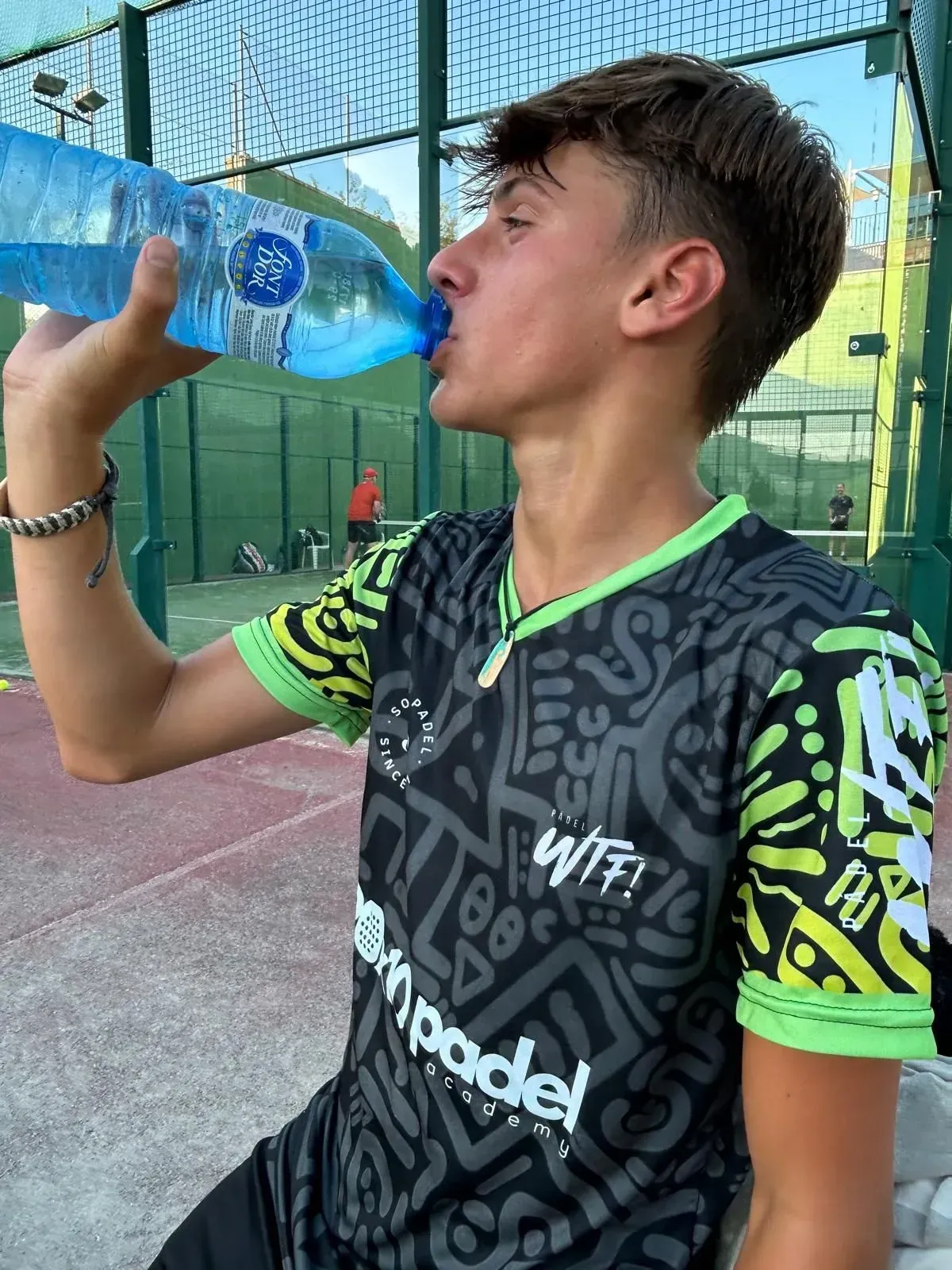 Un joven bebe agua de una botella mientras viste una camiseta de pádel.
