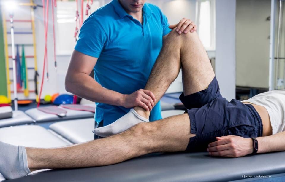 Physiotherapeut untersucht Knie und Knöchel des Patienten. Hellhäutig, blaues Hemd und Shorts.