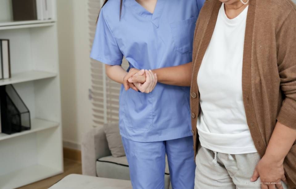 Krankenschwester im blauen Kittel hilft einer älteren Frau beim Gehen. Weiße und braune Kleidung in einer häuslichen Umgebung.