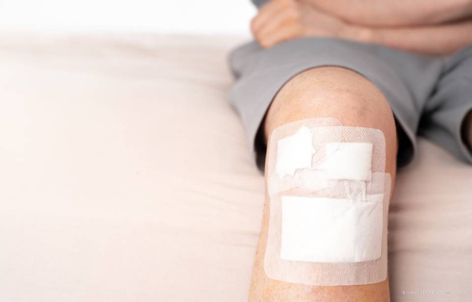 Knie mit zwei Bandagen, Person in grauen Shorts sitzt auf einer beigen Fläche.
