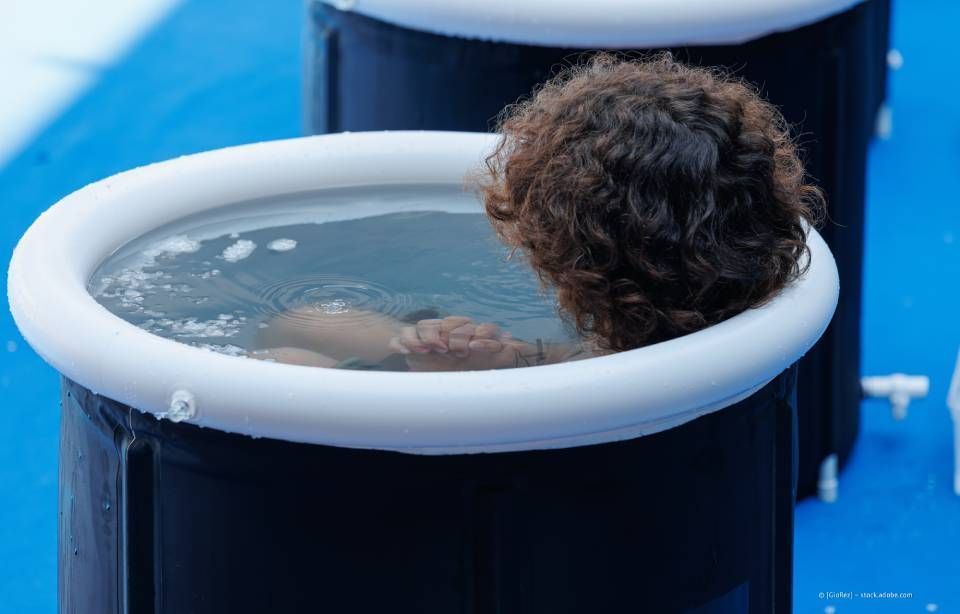 Person mit lockigem braunem Haar in einem kleinen kalten Tauchbecken. Dunkle Wanne, weißer Rand, blaue Oberfläche.