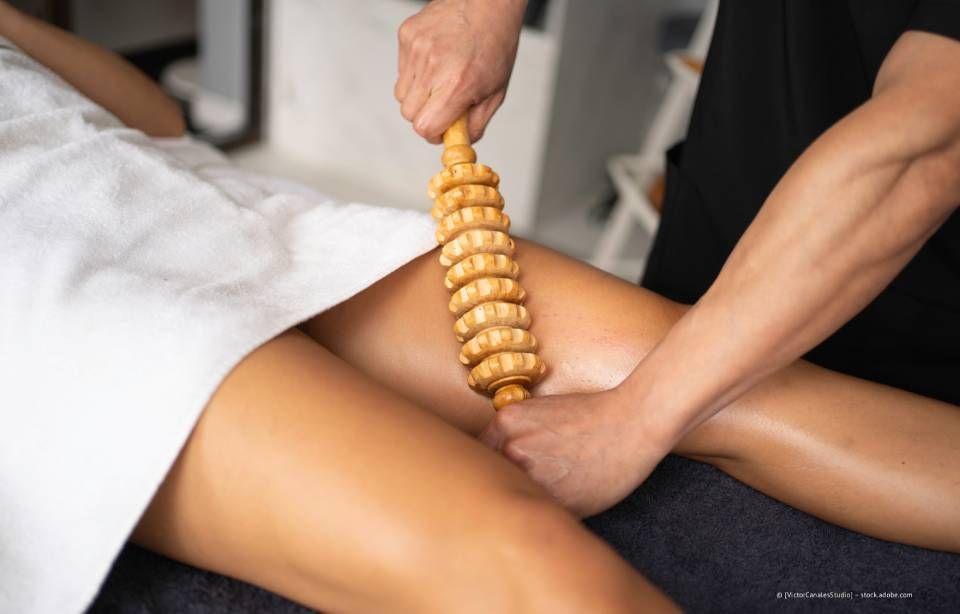 Therapeut verwendet ein hölzernes Massagegerät am Oberschenkel einer Person in einem Behandlungsraum.