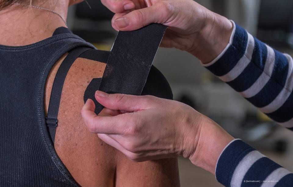 Person, der Kinesiologie-Tape an der Schulter angelegt wird; schwarzes Tape, Nahaufnahme.