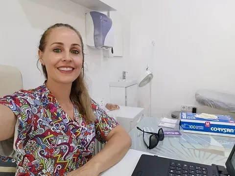 Mujer con uniforme colorido sonriendo a la cámara, sentada en un escritorio en un entorno médico.