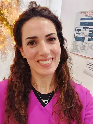 Una mujer con cabello castaño rizado sonríe, vistiendo una blusa médica y un collar rosa, parada cerca de una planta decorativa y un cartel.