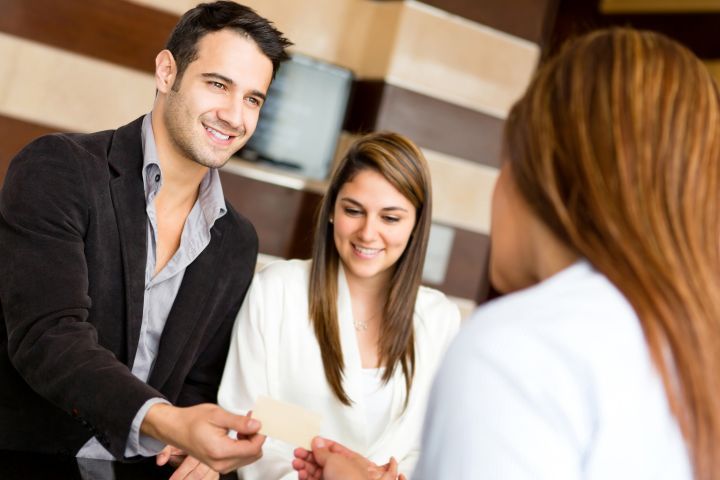 Mann überreicht einer Frau an der Hotelrezeption eine Karte. Frau und eine weitere Person lächeln.