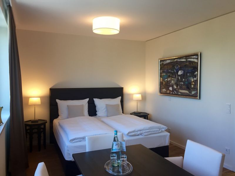 Schlafzimmer mit einem Bett, Nachttischen mit Lampen, Kunstwerken, Tisch mit Wasserflaschen und Stühlen.