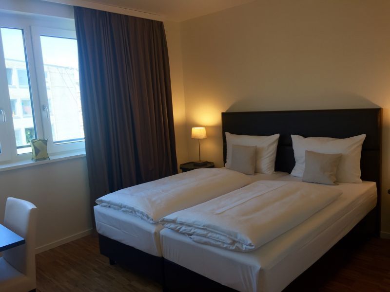 Schlafzimmer mit zwei Einzelbetten, weißer Bettwäsche, braunen Vorhängen, Fenster, Lampe auf dem Nachttisch und hellbraunen Wänden.