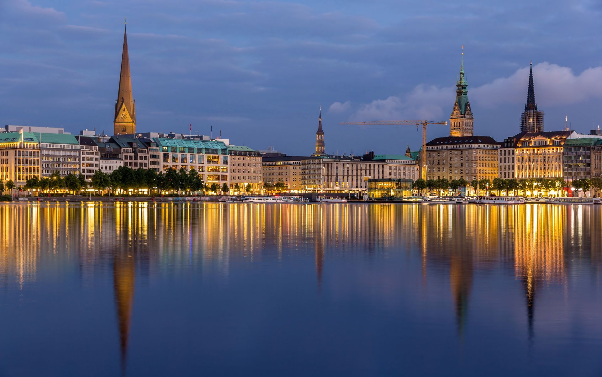 Hamburger Stadtbild in der Abenddämmerung, das sich im stillen Wasser spiegelt, mit beleuchteten Gebäuden und Kirchtürmen.
