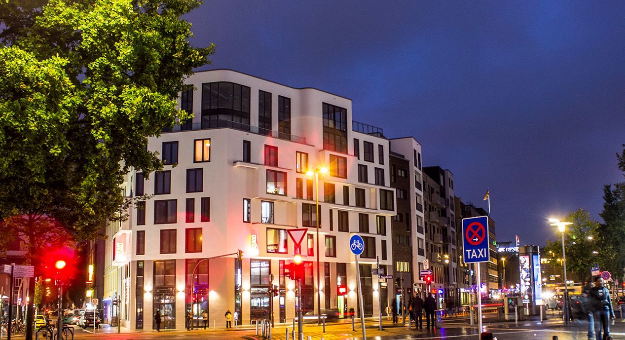 Weißes mehrstöckiges Gebäude mit beleuchteten Fenstern bei Nacht, Straße mit Ampeln und Fußgängern.