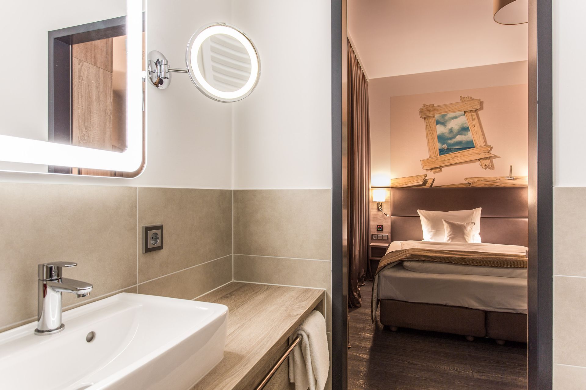 Badezimmer mit beleuchtetem Spiegel und Waschbecken, Tür zu einem Schlafzimmer mit Bett, Gemälde und Lampe.