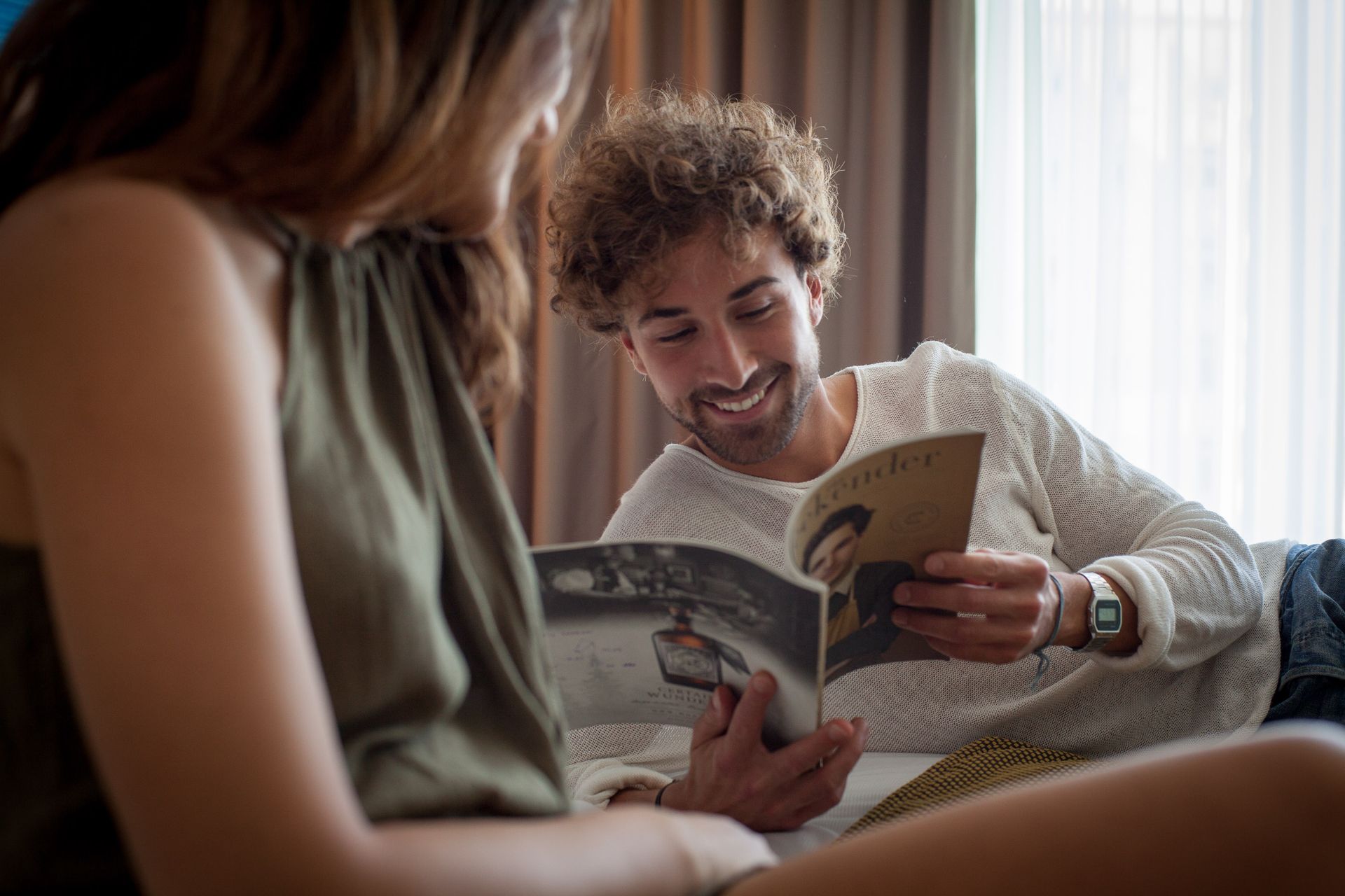 Mann mit lockigem Haar lächelt, während er eine Zeitschrift liest, neben einer Frau in einem Raum.