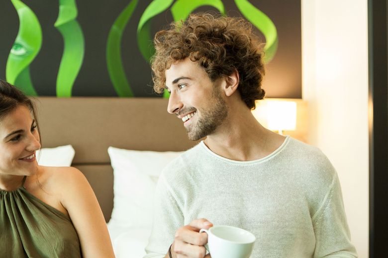Lächelnder Mann mit lockigem Haar und einer Tasse, der mit einer Frau in einem Schlafzimmer spricht.