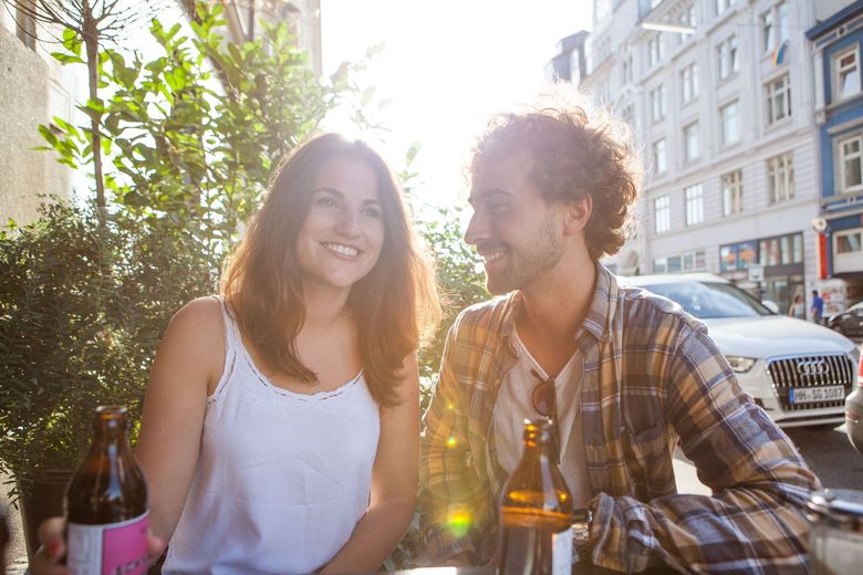 Lächelndes Paar in einem Straßencafé, von der Sonne beleuchtet. Frau in Weiß, Mann in Plaid, Flaschen auf dem Tisch.