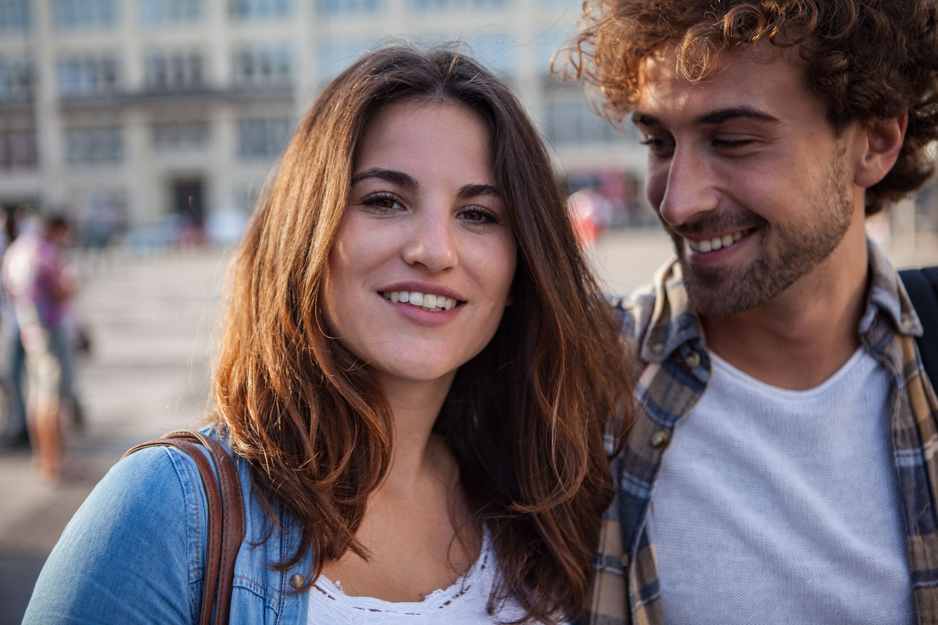 Frau mit braunem Haar lächelt in die Kamera, während ein Mann mit lockigem Haar zusieht, draußen in einer Stadt.