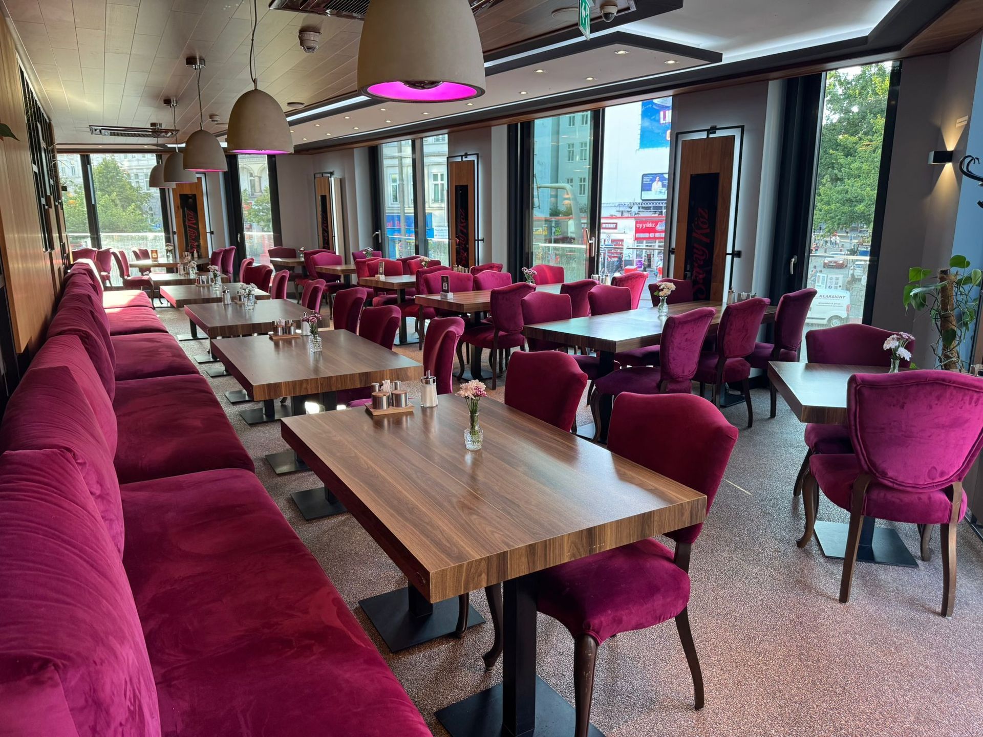 Restaurantinnenraum mit kastanienbraunen Sitzgelegenheiten, quadratischen Holztischen und großen Fenstern.