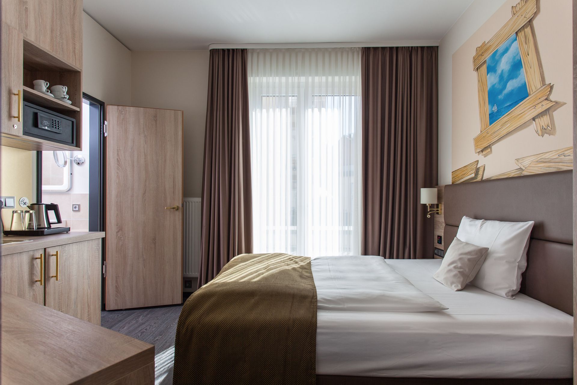 Hotelzimmer mit Bett, Fenster mit Vorhängen, Minikühlschrank und dekorativer Wandmalerei.