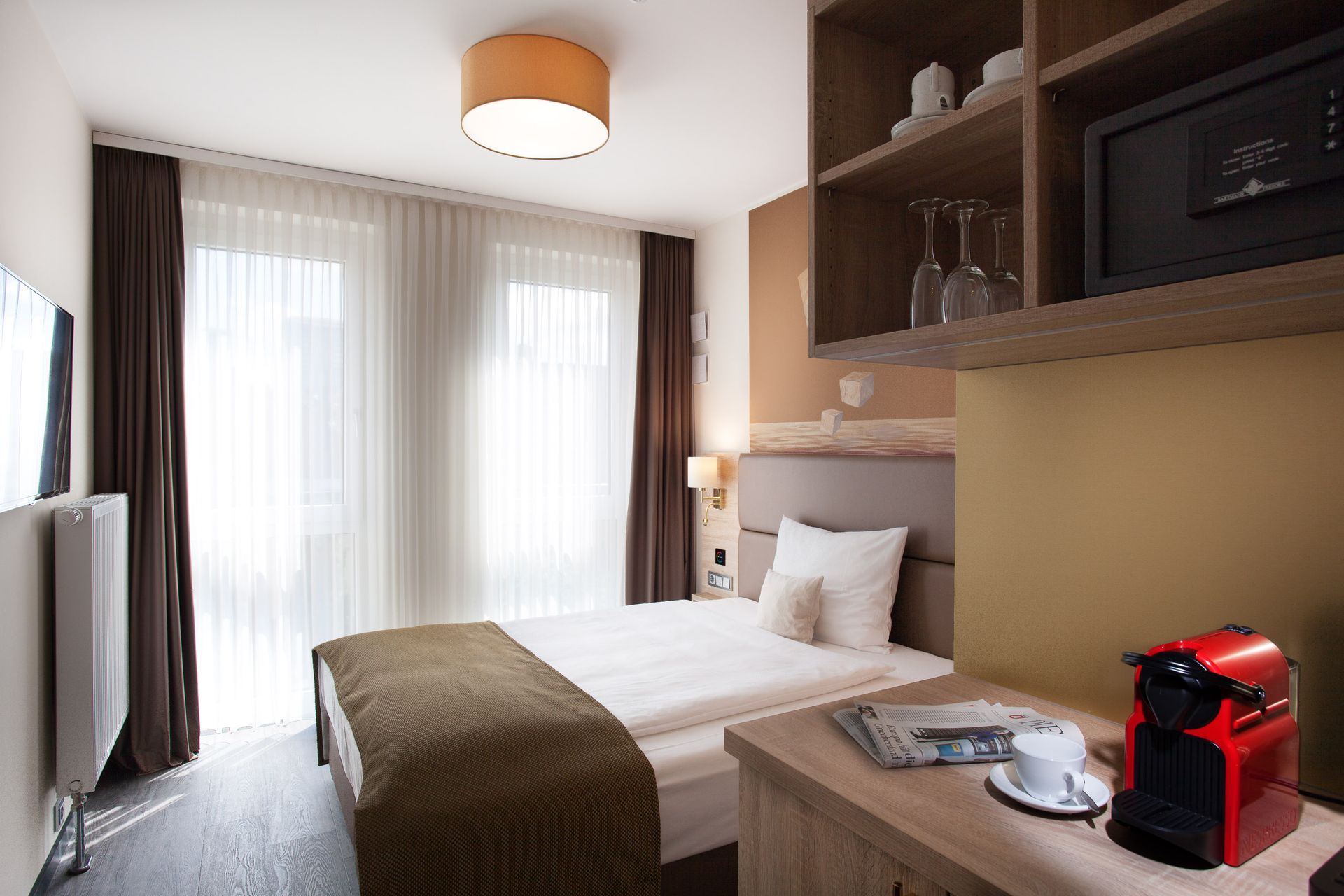 Hotelzimmer mit Bett, Schreibtisch mit Kaffeemaschine und großem Fenster mit transparenten Vorhängen.