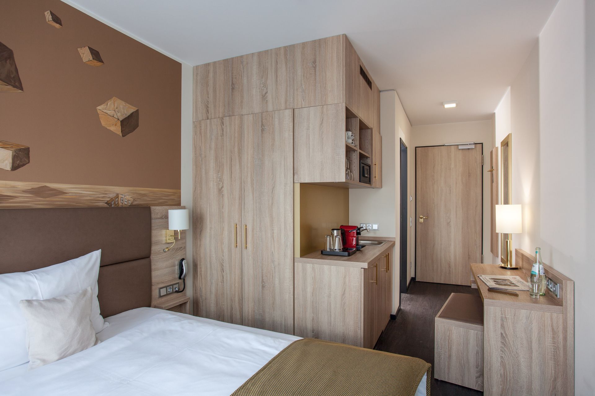 Hotelzimmer mit Bett, Schreibtisch und hellen Holzeinbauschränken und einer braunen Wand mit kunstvollen Würfeln.
