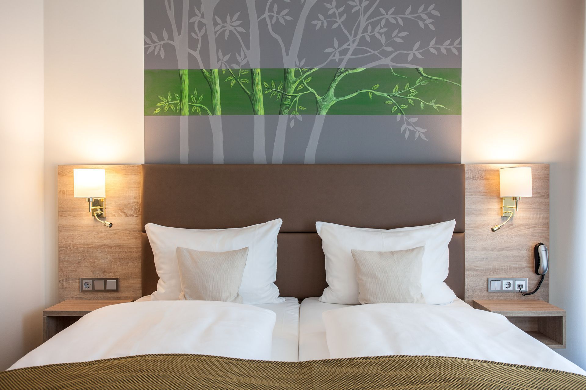 Hotelzimmer mit einem Doppelbett, zwei Lampen und einer Wanddekoration mit Baummotiven und grünen Akzenten.