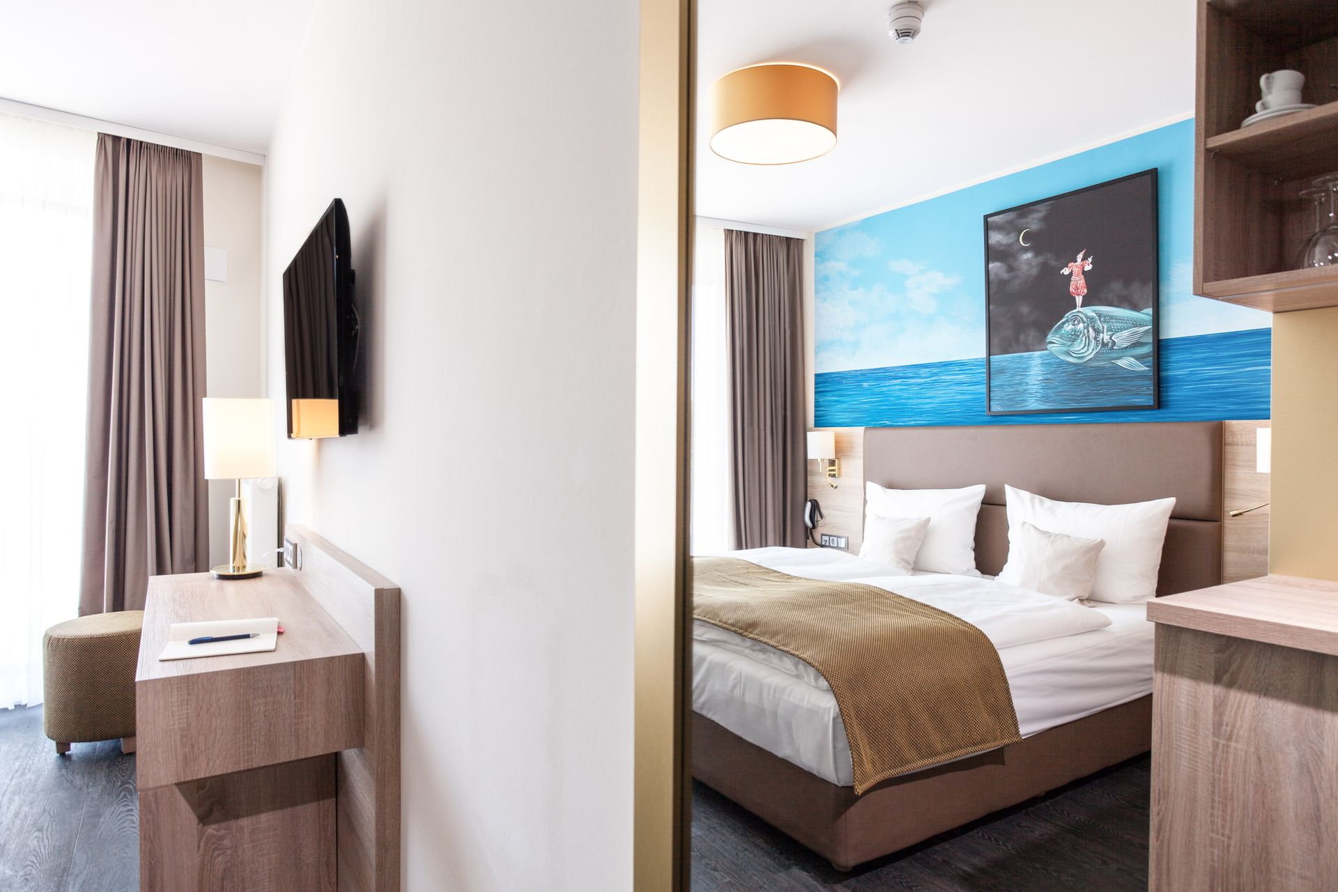 Hotelzimmer mit Bett, Kunstwerk, Fernseher, Schreibtisch und Fenster; Beige-, Blau- und Brauntöne.