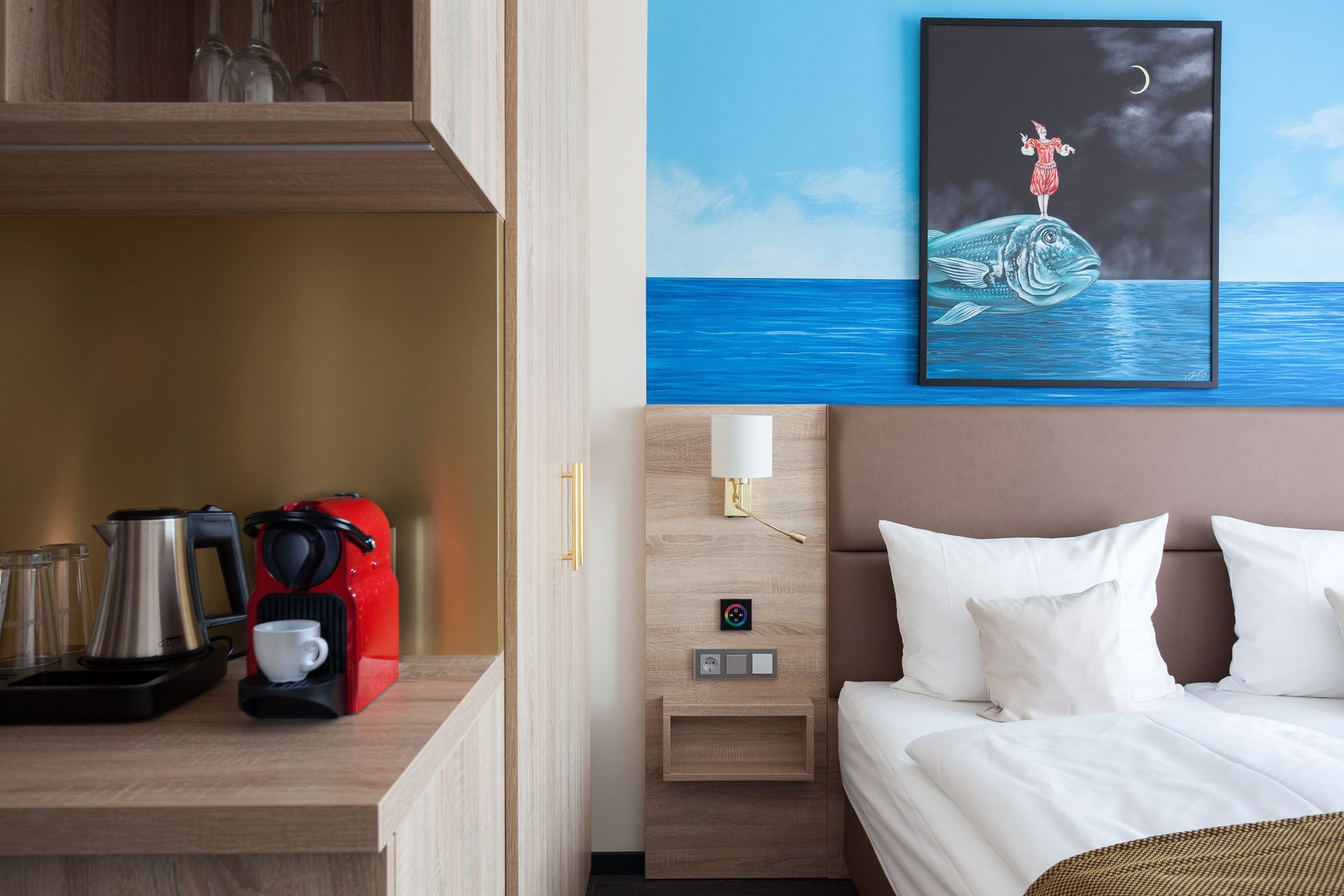 Hotelzimmer mit Bett, Kunstwerken, Kaffeemaschine und hellen Holzmöbeln.