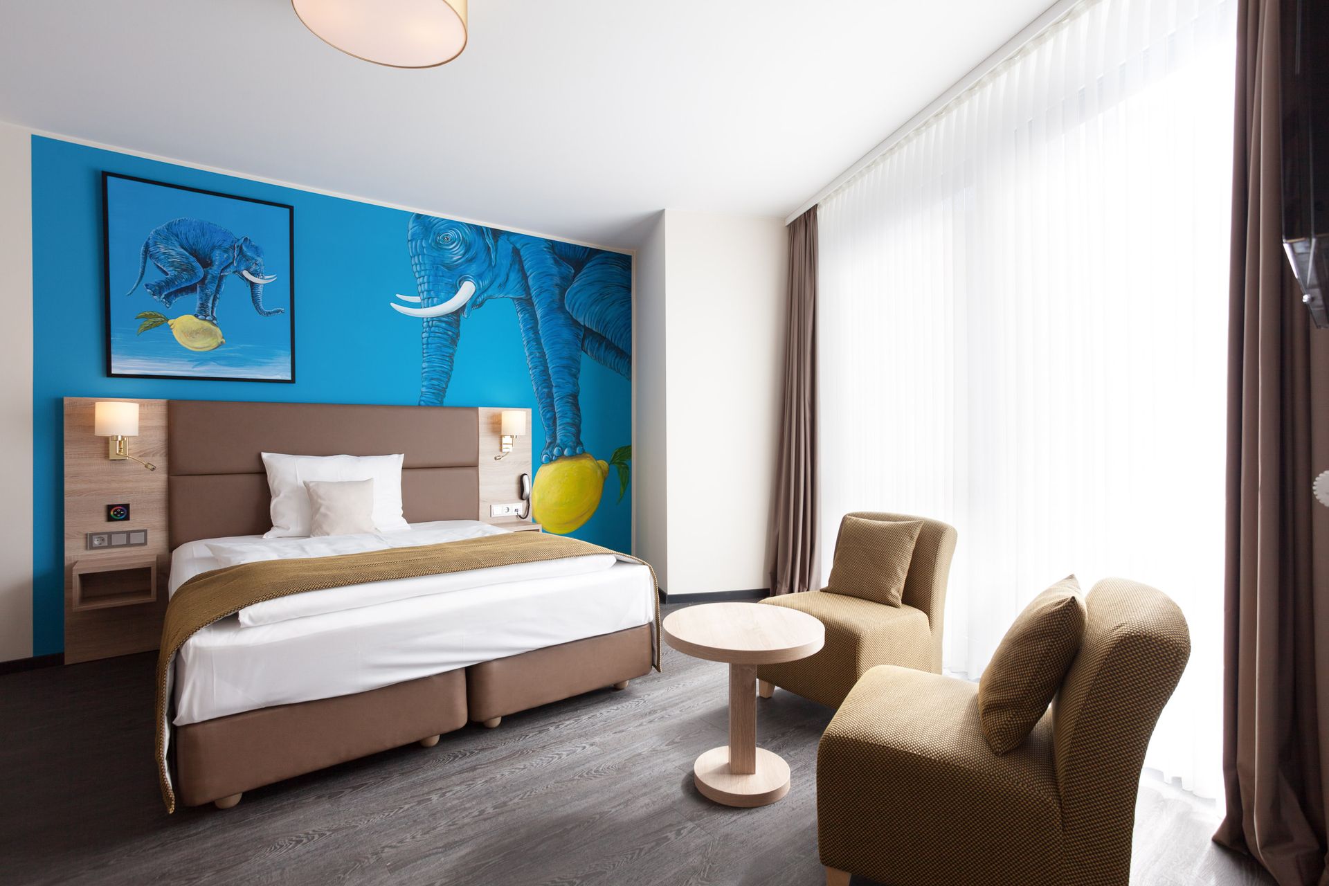 Hotelschlafzimmer mit blauer Akzentwand, Bett, zwei Stühlen, kleinem Tisch und großem Fenster.