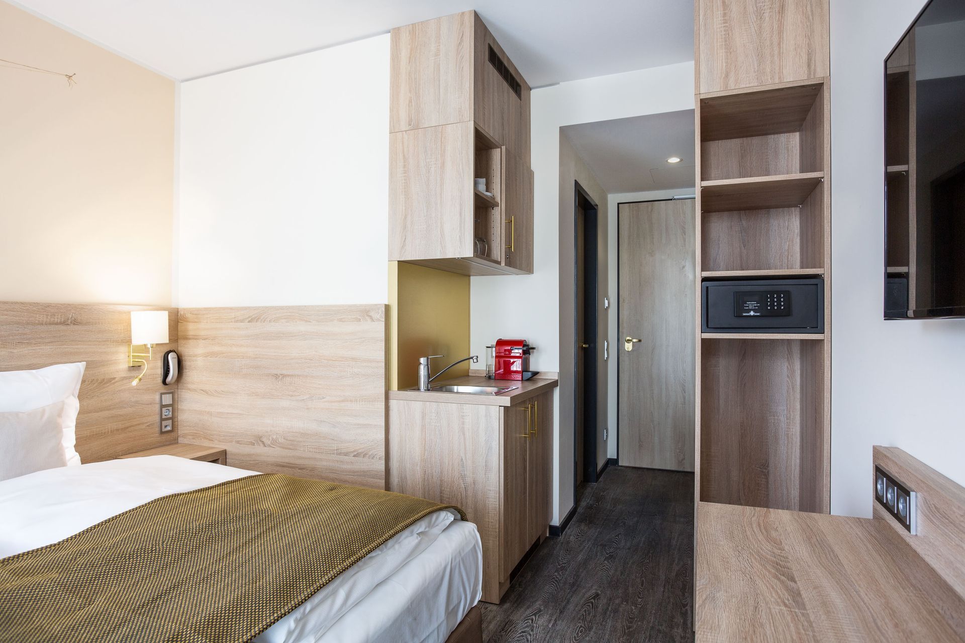 Kleines Hotelzimmer mit Bett, Waschbecken, Safe und Regalen. Beige- und Holztöne.