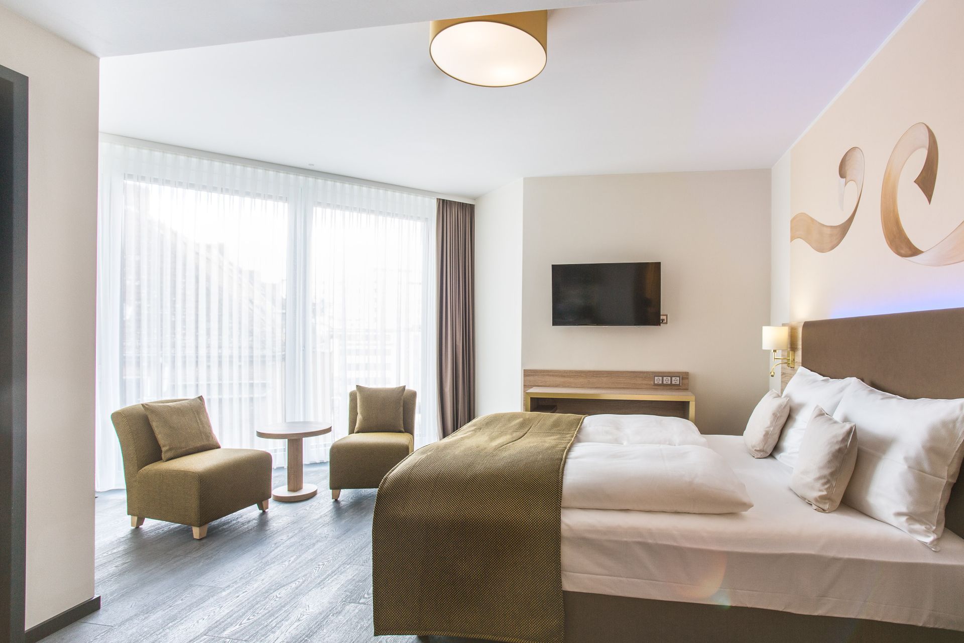 Hotelzimmer mit Bett, Stühlen, großem Fenster und Fernseher. Helle Wände, dunkler Boden und neutrale Farben.