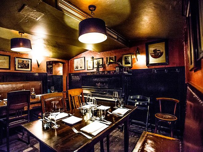 Dunkles, stimmungsvolles Interieur eines Restaurants mit einem langen Holztisch für die Gäste; gerahmte Kunst und ein Klavier sind zu sehen.