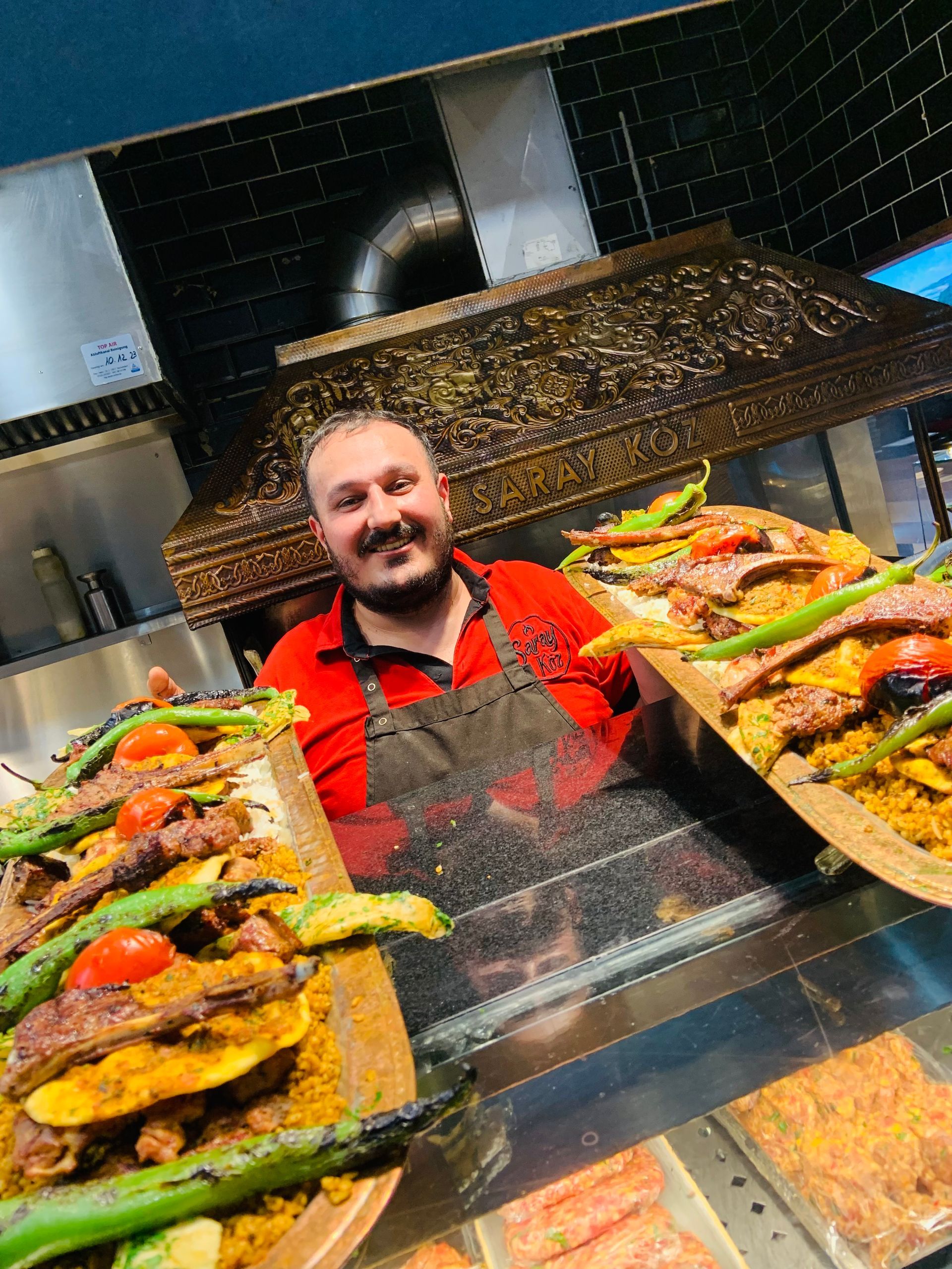 Mann in rotem Hemd und Schürze hält Platten mit Essen in einer Restaurantküche. Lächelnd, mit Essen auf der Schautafel.