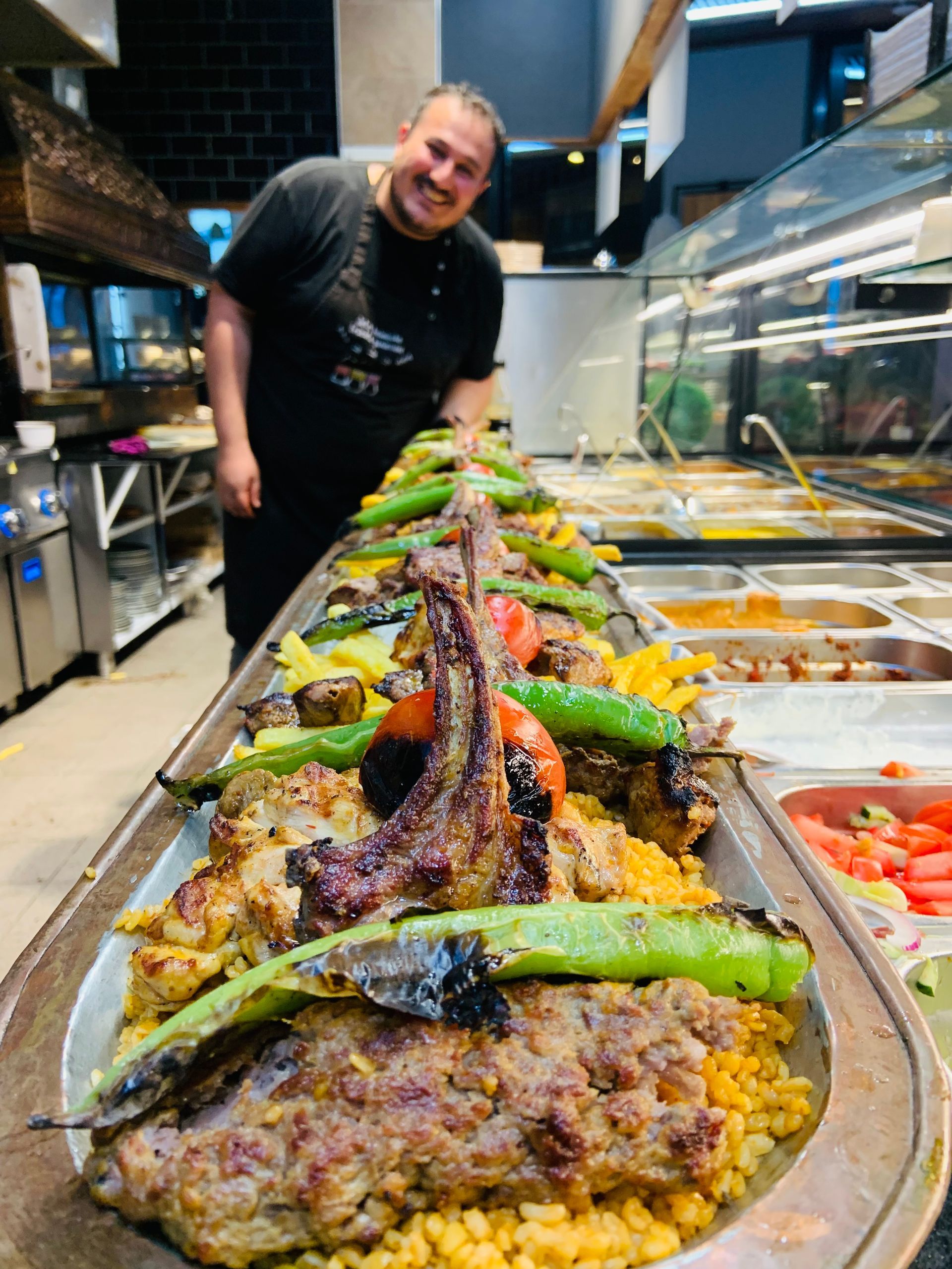 Der Koch lächelt hinter einem langen Tablett mit gegrilltem Fleisch, Gemüse und Reis in einem Restaurant.