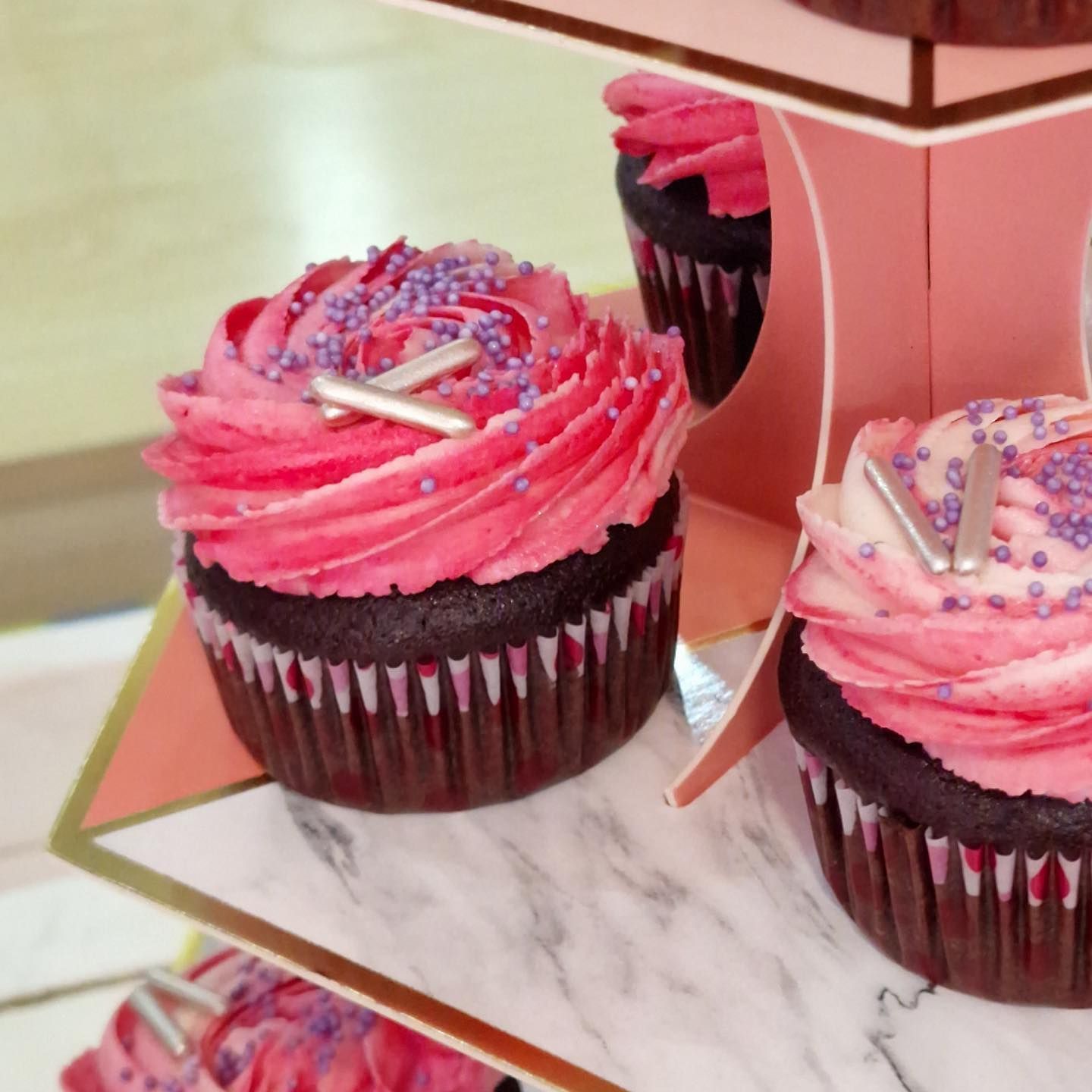 Cupcakes con glaseado rosa, chispas violetas y detalles plateados sobre un soporte escalonado de color rosa.