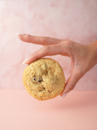 Mano sosteniendo una galleta redonda con chispas de chocolate sobre un fondo rosa.