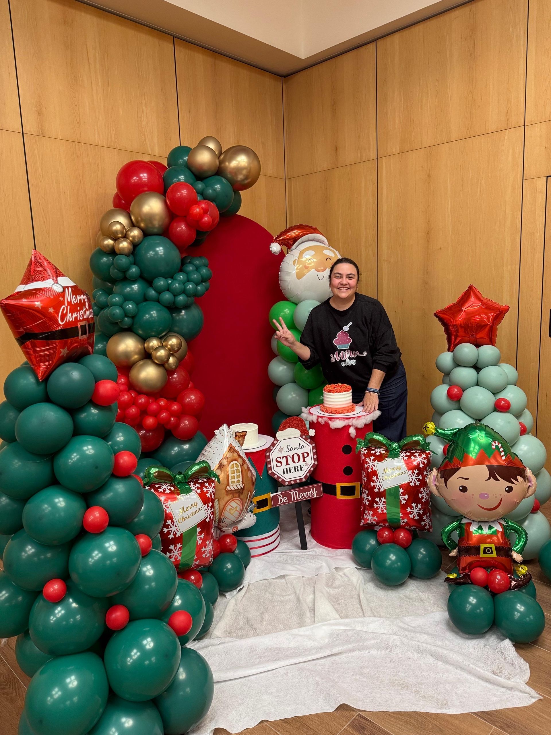 Mujer se encuentra en medio de decoraciones de globos navideños: árboles, Papá Noel, arco.