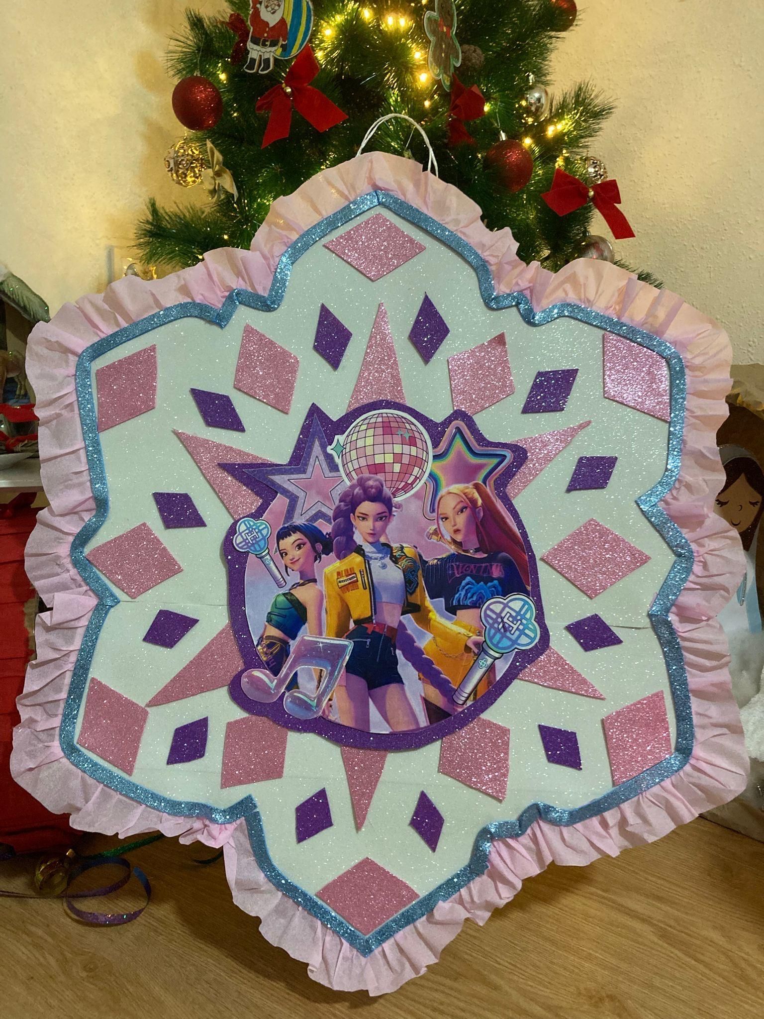 Piñata en forma de estrella con una foto grupal de Barbie, detalles de brillo rosa, morado y azul, bordes con volantes, frente a un árbol de Navidad.