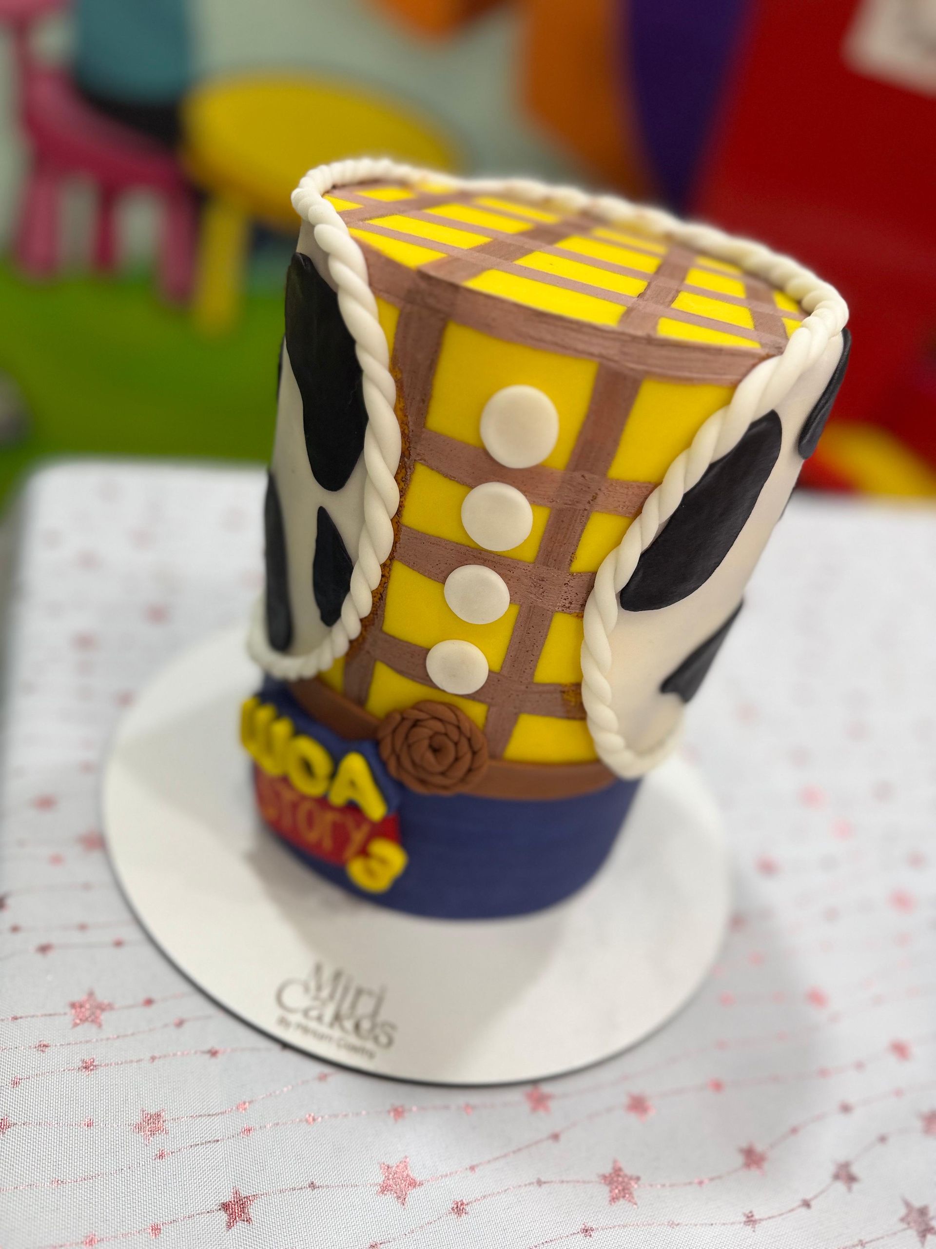 Pastel con sombrero de Woody de Toy Story, amarillo con patrón de cuadrícula marrón, lados con estampado de vaca, borde de cuerda blanca, en plato blanco.
