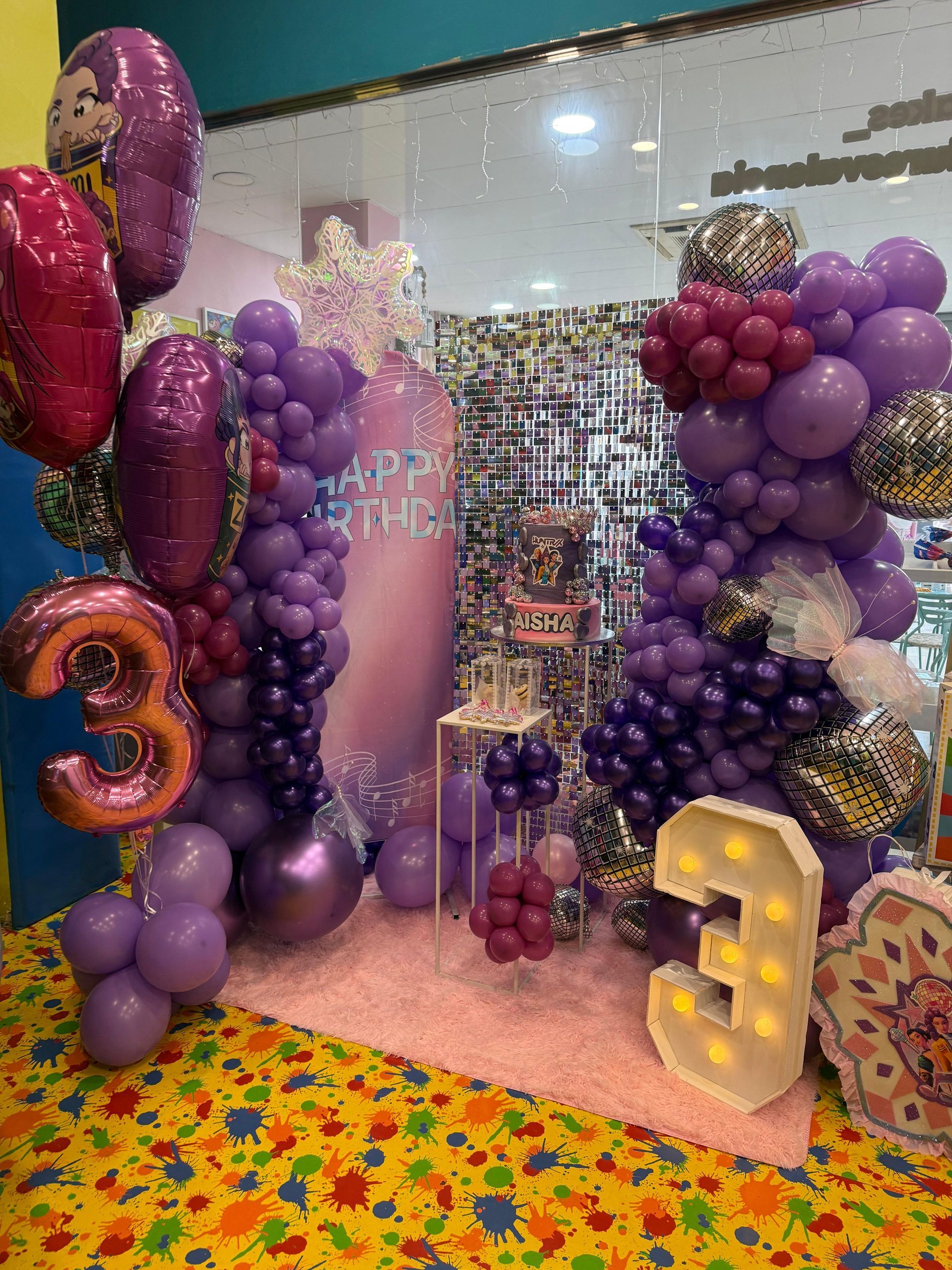 Una fiesta de cumpleaños preparada con globos morados y rosas, un pastel y el número 