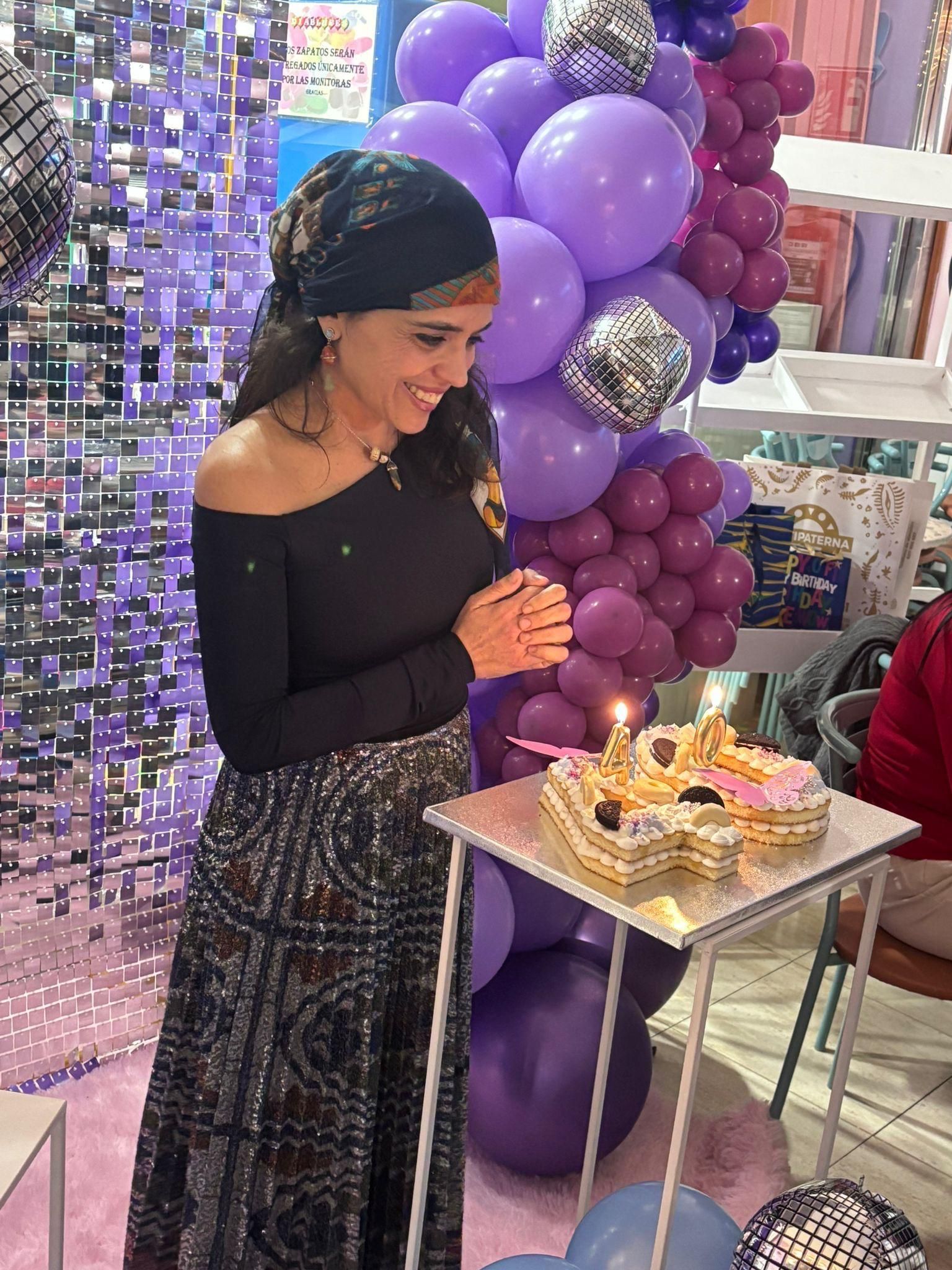 Mujer con las manos juntas, sonríe ante un pastel en forma de corazón con vela, globos morados de fondo.