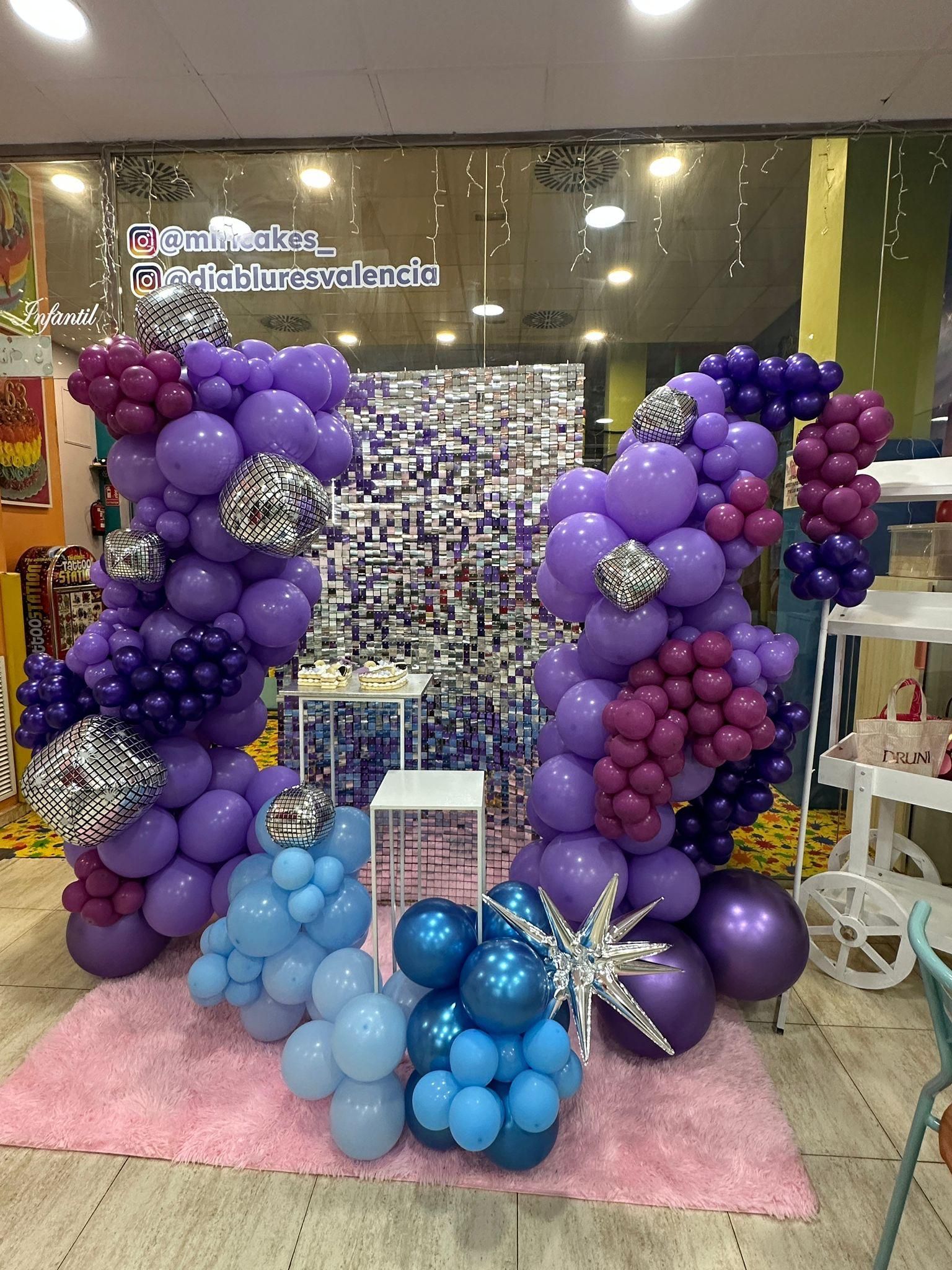 Decoración de arco de globos morados y azules con bolas de discoteca sobre alfombra rosa.