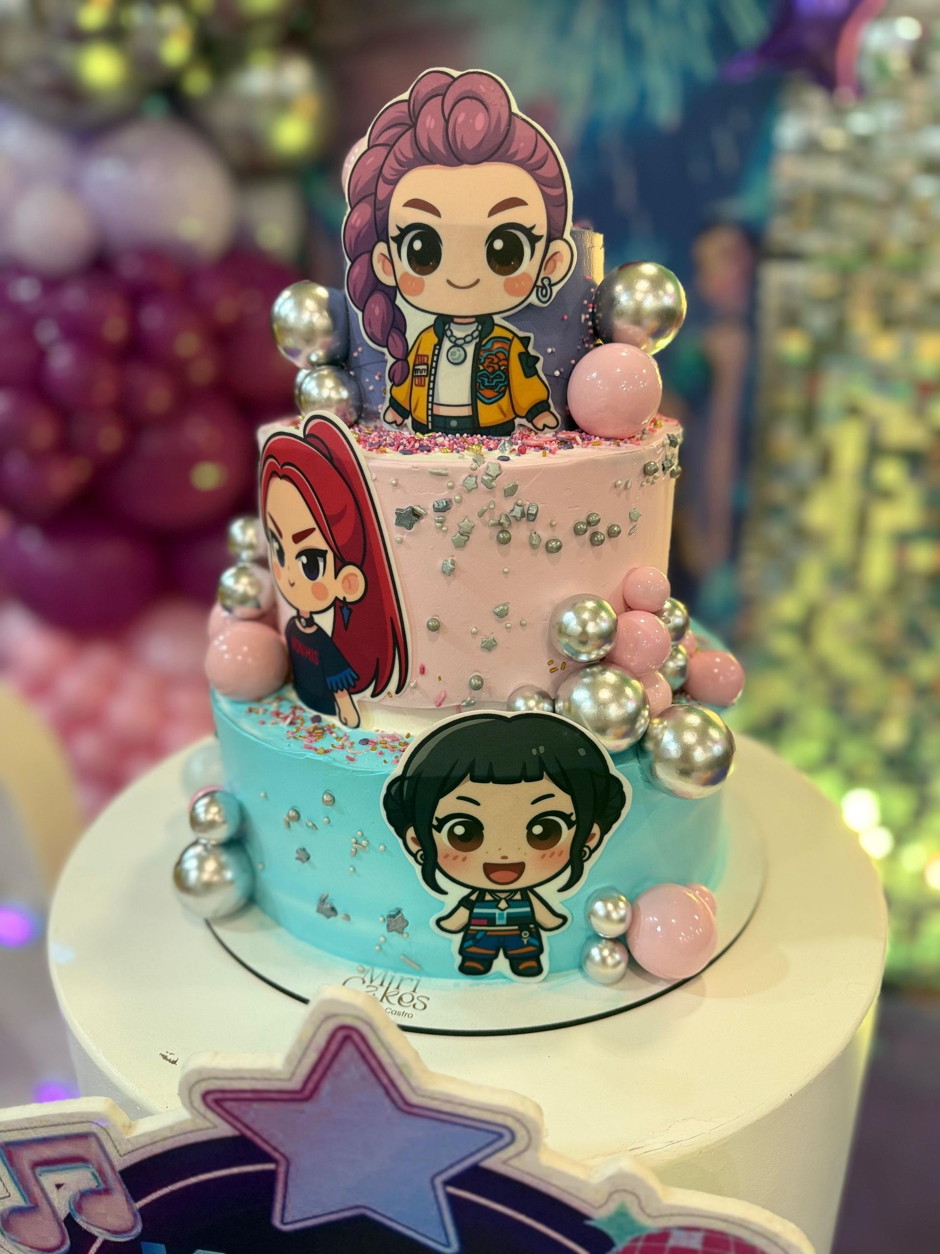 Pastel de cumpleaños de tres pisos con adornos de los personajes de K/DA. Glaseado rosa y verde azulado, chispas plateadas y detalles perlados.