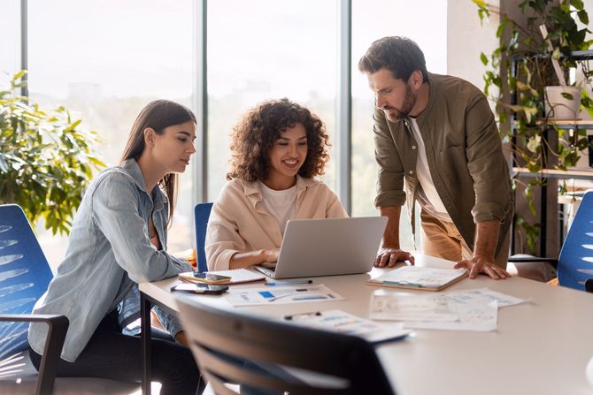Trois personnes collaborent autour d'un ordinateur portable et de documents, assises à une table dans un bureau lumineux.