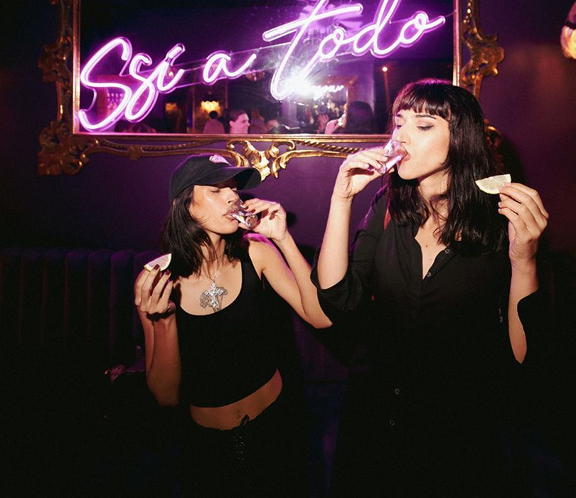Dos mujeres bebiendo frente a un letrero de neón que dice sí a todo