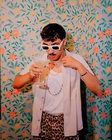 Un hombre que lleva gafas de sol sostiene una copa de martini frente a una pared floral.